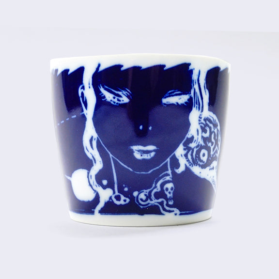 White ceramic cup with a wrap around dark blue illustration. Current side features a close up of a woman's face with a bird's head peeping over her shoulder.