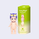 Vinyl figure of a Kewpie baby standing nude with a purple owl hood hat atop its head. It stands next to a pink display box that reads "Sonny Angel Animal Series Ver. 1"