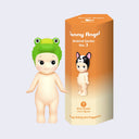 Vinyl Kewpie baby figure, standing nude while wearing a green crocodile head as a hooded hat. Beside is its display box, orange to cream ombre with a small Kewpie baby wearing a French Bulldog head for a hat.