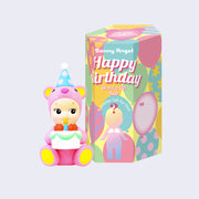 Vinyl figure of a Kewpie baby in a pink teddy bear costume, with a blue party hat and holding a cake. It sits next to its product packaging.