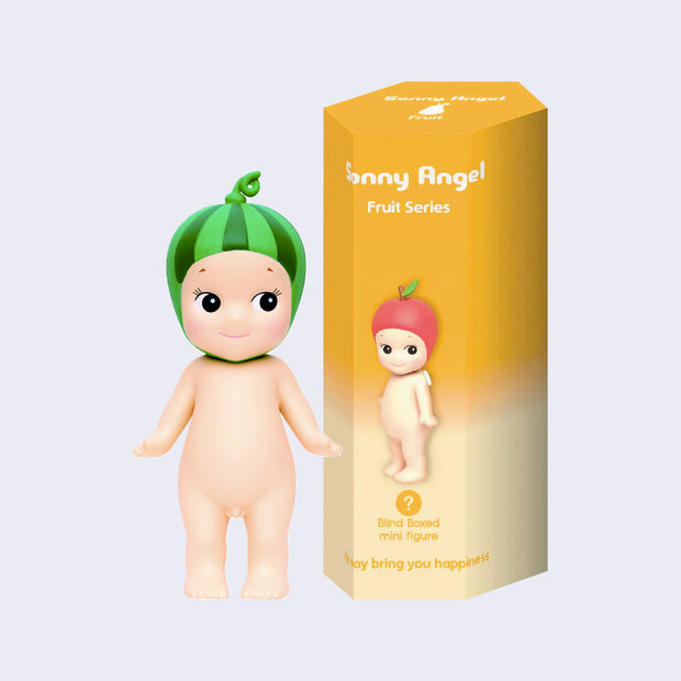 Vinyl Kewpie baby figure, standing nude while wearing a watermelon hat on its head. Beside is its display box, yellow to cream ombre with a small Kewpie baby wearing an apple hat.