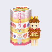 Small vinyl figure of a Kewpie baby wearing a large brown decorated cake as an outfit with strawberries too. It says "Happy Birthday" on it. Atop the Sonny Angel's head is a swirled hat of matching colored frosting with candles on top.