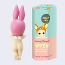 A small Kewpie baby figure, facing away so only their nude body is showing, their legs slightly crossed with a small pair of angel wings on their back. They wear a pink bunny hood hat. Next to them is a green and peach color display box with an illustration of a smiling Kewpie baby with a deer hood hat resting its arms flat. 