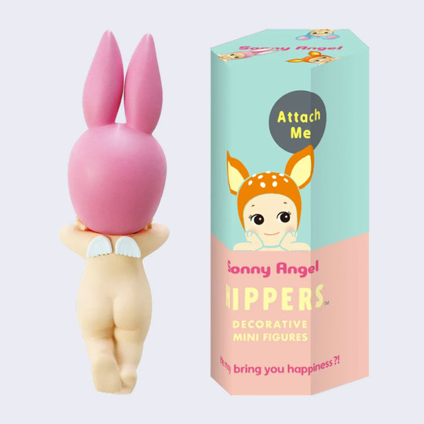 A small Kewpie baby figure, facing away so only their nude body is showing, their legs slightly crossed with a small pair of angel wings on their back. They wear a pink bunny hood hat. Next to them is a green and peach color display box with an illustration of a smiling Kewpie baby with a deer hood hat resting its arms flat. 