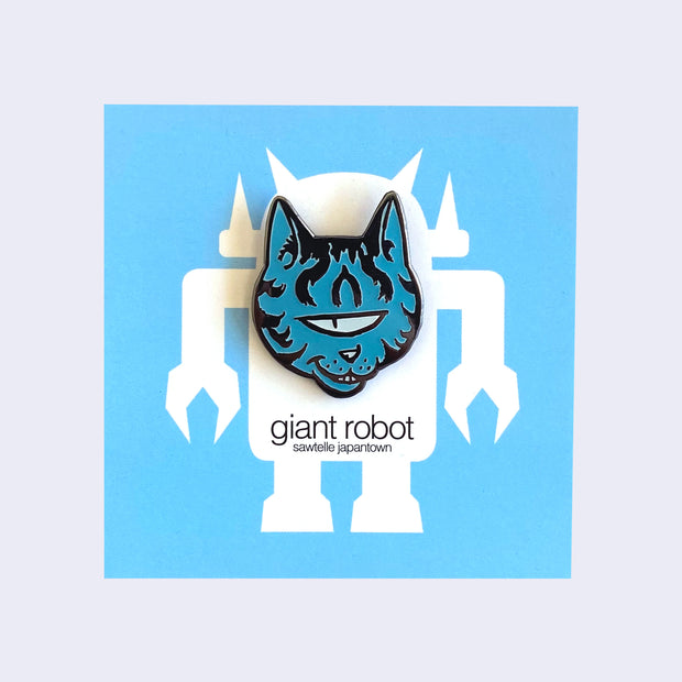 Enamel pin of blue, one-eyed cat head with sneaky expression and smile. Cat has metallic stripes on head and cheeks. Pin is on blue backing card.