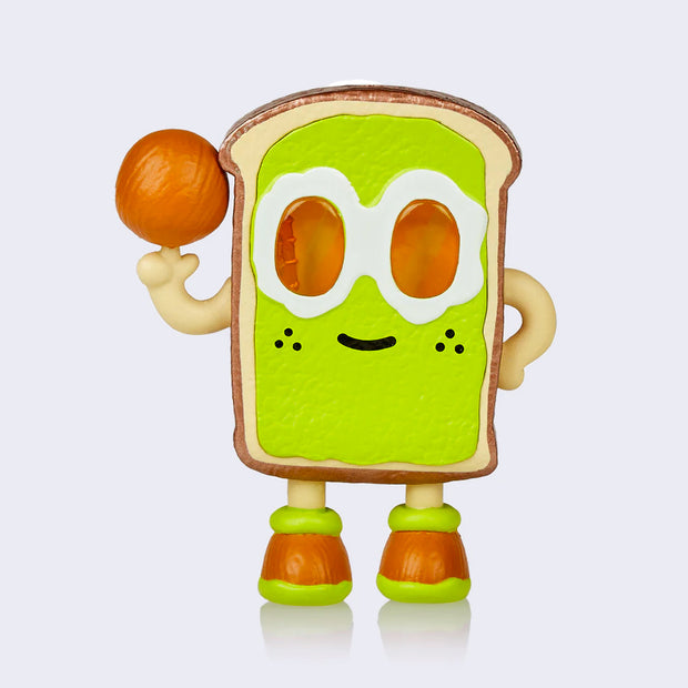 Vinyl figure of a slice of avocado toast, standing on 2 feet with sunny side eggs for eyes and a small smile. It poses like its spinning a basketball, but with an avocado pit.