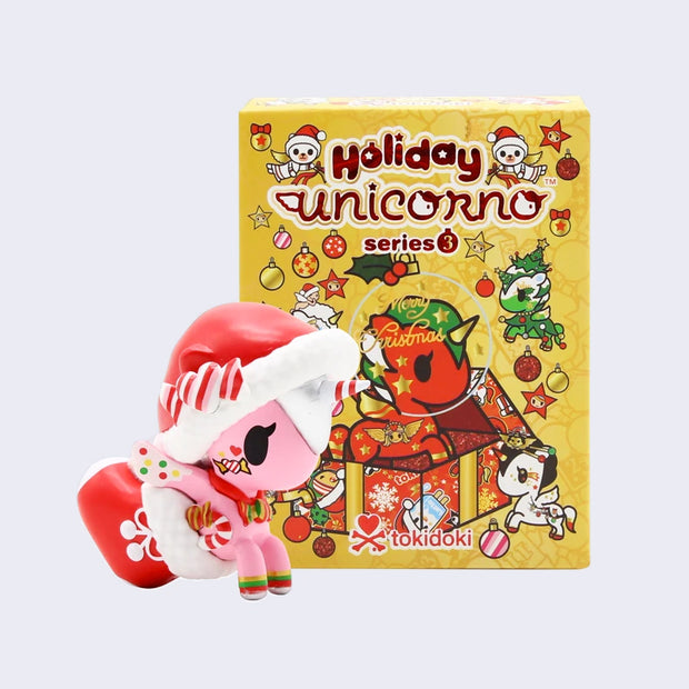 Vinyl pink unicorn figure, wearing a Santa hat with a candy cane heart on it. Its legs are inside of a red stocking with a white bow. It stands in front of a yellow blind box.