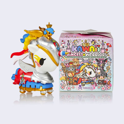 Vinyl unicorn figure, white with silver armor and red, blue and gold color details as if a warrior horse. Atop its head is a small golden baby unicorn with large wings. It stands next to its display box, colorful and full of tokidoki illustrations that reads "Kawaii Princess Warriors"