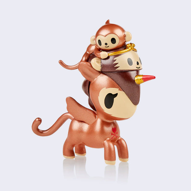 Vinyl metallic brown unicorn figure, with a lighter tan face and monkey tail. It wears a monkey head with a golden tiara on its head and a small smiling brown metallic monkey is grasping onto the hat.