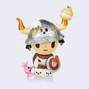 Mozzarella figure, a human in a cow costume with large gold tipped horns. She wears a silver helmet with a skull on it and a cracked egg. She wears fur armor, has a wooden shield with a burst strawberry on it, and a wrapped candy mallet.