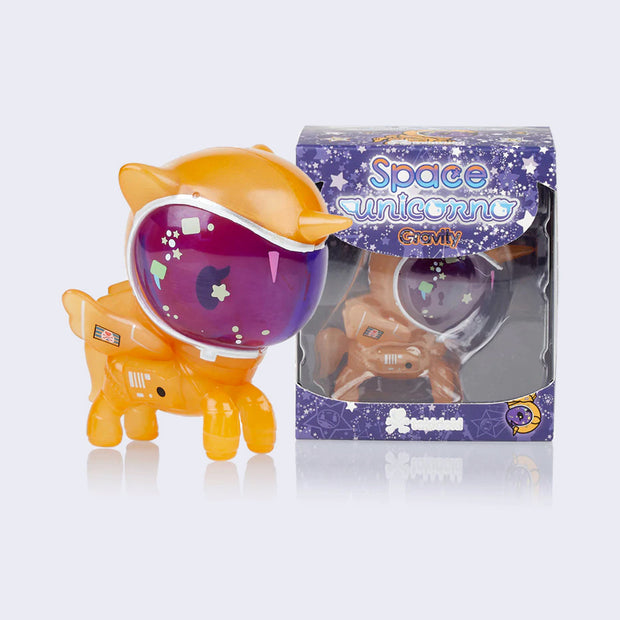 Vinyl unicorn figure wearing an orange space suit with a purple tinted face shield. Some stars and sparkles are appliquéd onto the face. It stands next to its clear display box packaging.