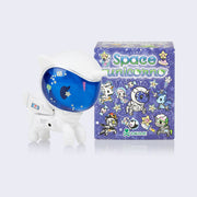 Vinyl unicorn figure, wearing a full body white space suit with a blue tinted face shield part of the helmet with stars and sparkles appliquéd on. It stands next to its blind box packaging, purple with illustrations of space themed unicorns.