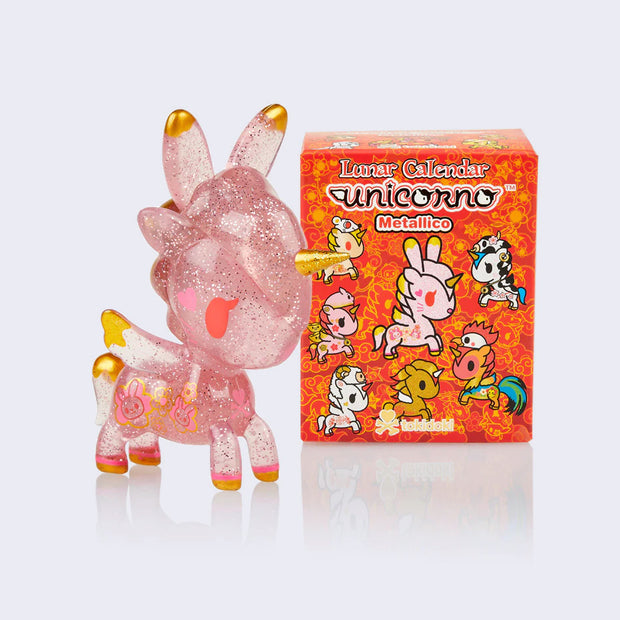 Vinyl pink transparent unicorn figure with glitter and bunny ears and appliqué bunnies and hearts on the side of its body. The tips of its ears, wings, tail, feet and horn all have gold accent coloring. It stands next to its blind box packaging, red with various illustrations of Unicornos dressed as different lunar calendar animals.