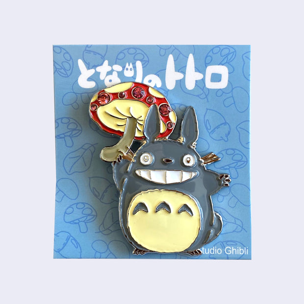 Brooch of Totoro smiling and holding a large red polka dot mushroom like an umbrella. Mushroom has 7 red gems on it.