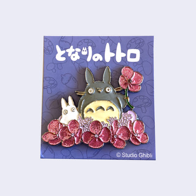 Brooch of Totoro and a small, white chibi Totoro in a field of glittery pink flowers. Totoro holds up a pink flower by its stem.