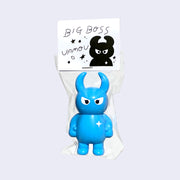 Cyan colored vinyl figure character, standing with arms at its side. It has a round head with curved horns and simple angry eyes with no other facial features. It has a white sparkle on its upper right chest. Character is inside of a plastic bag with a white hang tag that says "Big Boss Uamou" with an illustration of the figure.