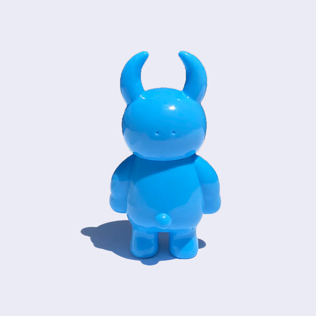 Back view of cyan colored vinyl figure character, standing with arms at its side. It has a round head with curved horns and a small spherical bump for a  tail.