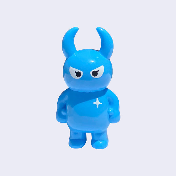 Cyan colored vinyl figure character, standing with arms at its side. It has a round head with curved horns and simple angry eyes with no other facial features. It has a white sparkle on its upper right chest.