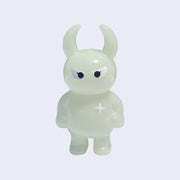 Light yellow/green colored vinyl figure character, standing with arms at its side. It has a round head with curved horns and simple angry eyes with no other facial features. It has a white sparkle on its upper right chest.