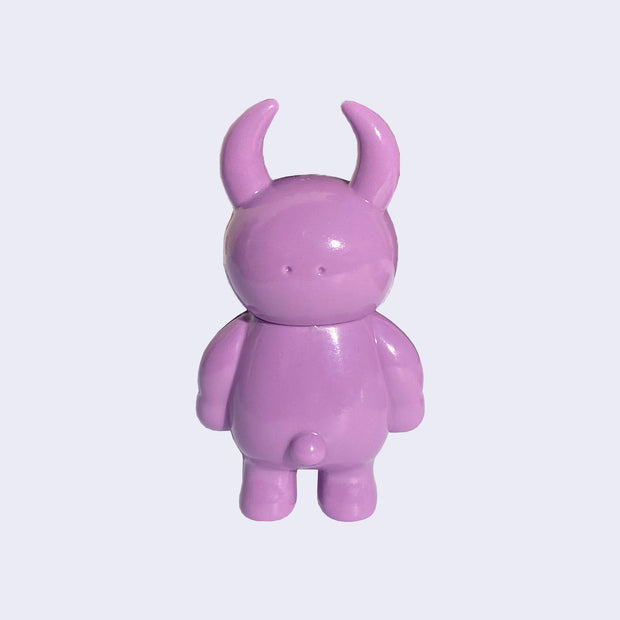 Back view of mauve colored vinyl figure character, standing with arms at its side. It has a round head with curved horns and a small spherical bump for a tail.