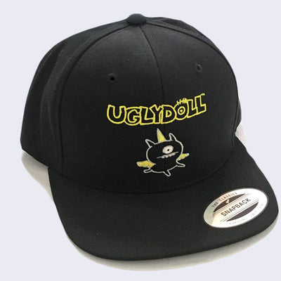 Front side of black hat. Stitched on front is a one-eyed monster with wings. Above the monster is yellow stylized text that says ugly doll.