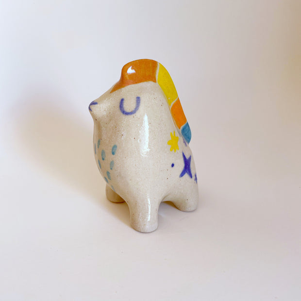 Small ceramic of an abstract figure, sitting on all fours with its eyes closed and a mane that runs down its head and back of orange, yellow and blue. It has colorful sparkled on its side.