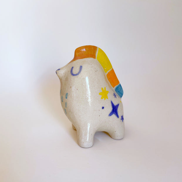 Small ceramic of an abstract figure, sitting on all fours with its eyes closed and a mane that runs down its head and back of orange, yellow and blue. It has colorful sparkled on its side.