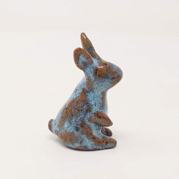Brown and blue ceramic simplistic shaped bunny, without any distinct facial features. Its ears are up and one paw is raised, curiously.