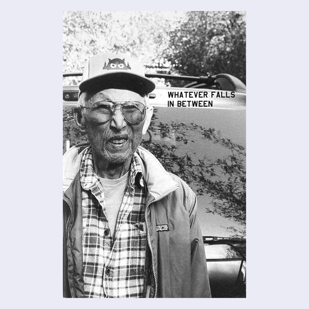 Black and white zine cover featuring a photograph of an older man wearing in front of a car rear. "Whatever Falls In Between" is written in thin, all caps font.