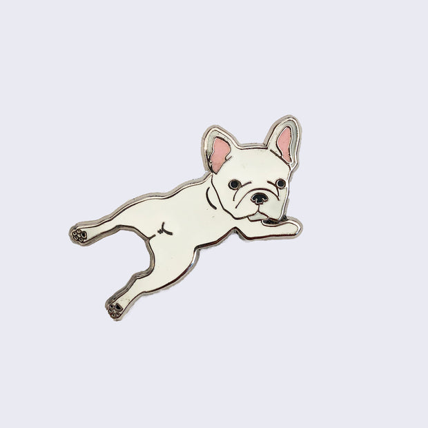 Enamel pin of a cream colored French Bulldog, laying on its stomach and its head turned to look straight at the viewer.