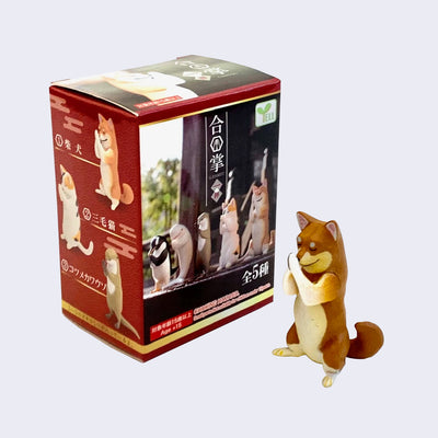 Small plastic brown and white Shiba Inu figure, with its head bowed slightly, eyes closed and little hands touching in prayer. It stands next to its blind box packaging, red with bowing plastic shiba, calico cat and otter.