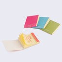 Set of 4 blocks of yellow sticky notes, each with a colored paper outer cover like a matchbook. Colors are: pink, blue, green and orange.