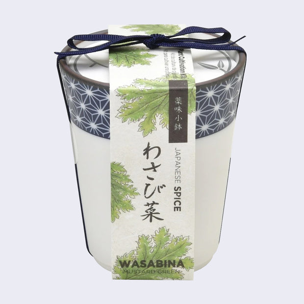 A white ceramic cup with blue dot patterning around the rim and a paper label that reads "Wasabina - Mustard Green" with illustrations of leaves. A blue string bundles the label and the cup.
